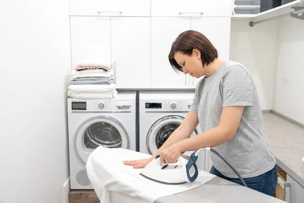 Молода жінка прасує білу сорочку на борту в пральній кімнаті з пральною машиною на фоні — стокове фото