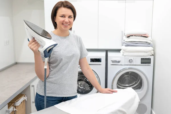 Mujer sonriendo mientras está de pie cerca de tabla de planchar en la sala de lavandería con lavadora en el fondo — Foto de Stock