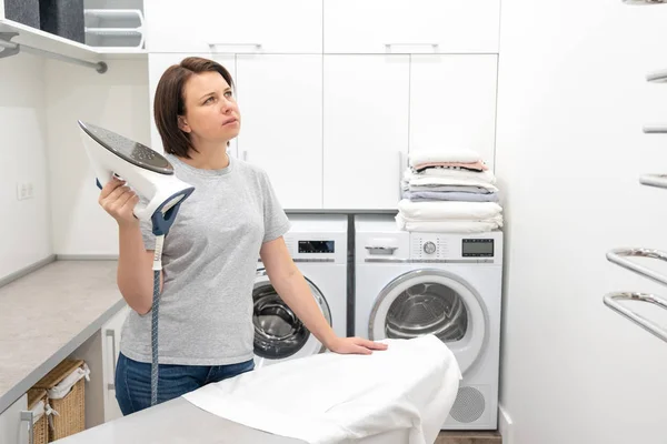 Жінка джакує під час прасування білої сорочки на борту в пральній кімнаті з пральною машиною на фоні — стокове фото