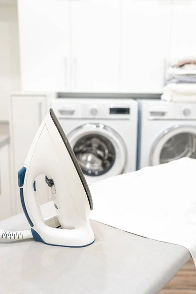 Besi di papan setrika dengan kemeja putih di ruang cuci — Stok Foto