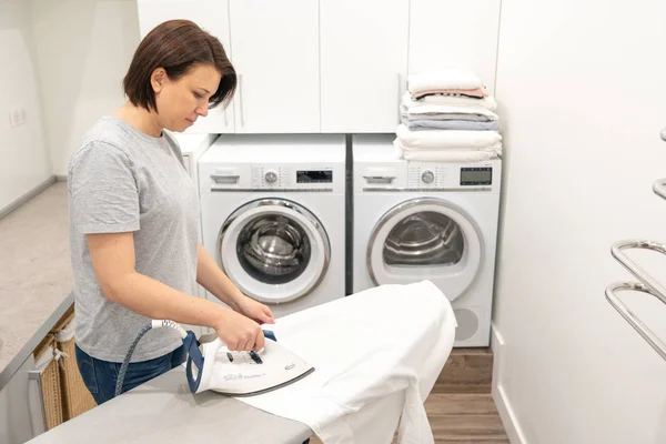 Woman ironing white clothes on board in laundry room with washing machine on background — Stock Photo, Image