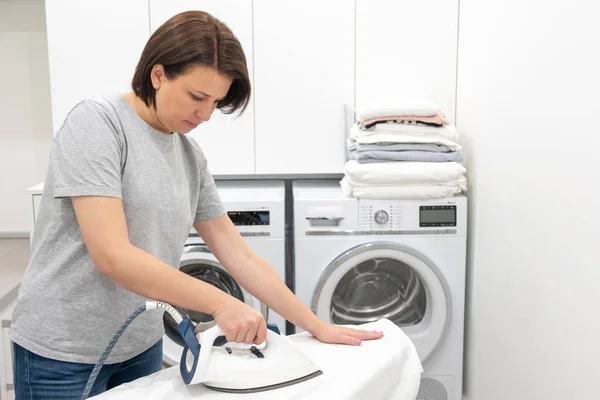 Woman ironing on board in laundry room with washing machine on background