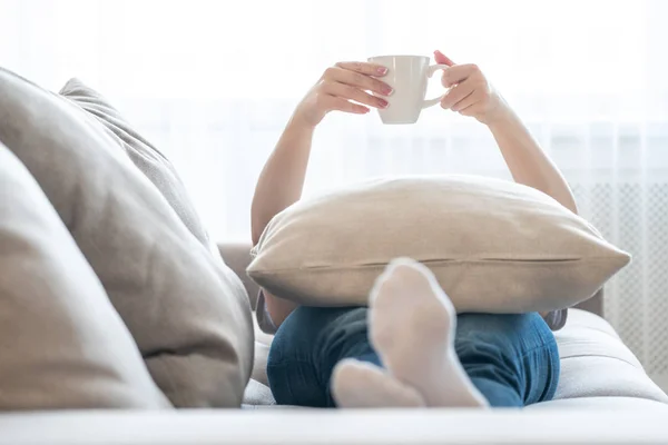 Genç bir kadın yastıkta bir fincan kahveyle kanepede dinleniyor. — Stok fotoğraf