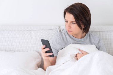 Yatağında yorganla oturan akıllı telefon kullanan bir kadın.