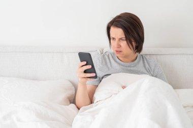 Yatağında yorganla otururken akıllı telefon kullanan sürpriz bir kadın.