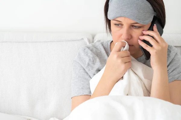 Mulher triste usando máscara de dormir usando smartphone como ela se senta na cama coberta com edredão — Fotografia de Stock