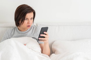 Yatağında yorganla otururken akıllı telefon kullanan sürpriz bir kadın.