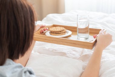 Elinde diyet kahvaltısı olan bir kadın yatakta kraker ve su tutuyor.