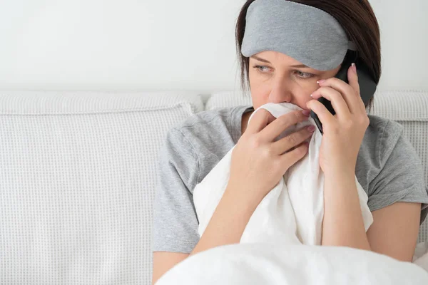 Donna triste che indossa maschera addormentata utilizzando smartphone mentre si siede nel letto coperto di piumone — Foto Stock