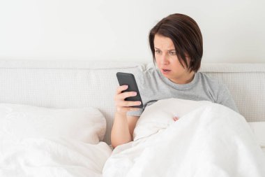 Yatağında yorganla otururken akıllı telefon kullanan sürpriz bir kadın.