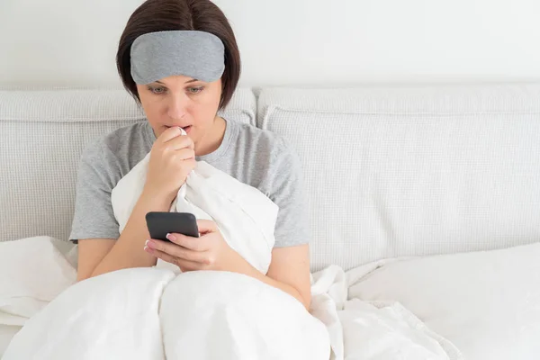 Kobieta w masce do spania smsuje na smartfonie, gdy siedzi w łóżku pokryta kołdrą — Zdjęcie stockowe