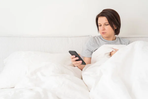Yatağında yorganla oturan akıllı telefon kullanan bir kadın. — Stok fotoğraf