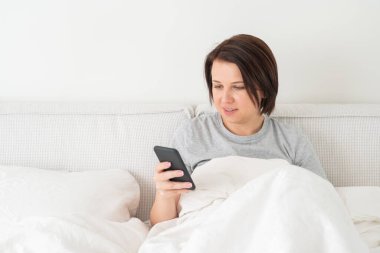 Yatağında yorganla oturan akıllı telefon kullanan bir kadın.