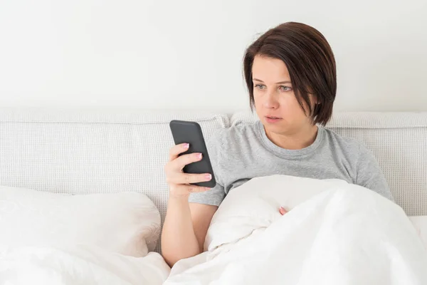 Yatağında yorganla otururken akıllı telefon kullanan sürpriz bir kadın. — Stok fotoğraf