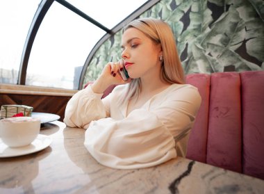 Genç bir kadın sıcak kahve içiyor ve kaldırımdaki kafede telefonla konuşuyor.