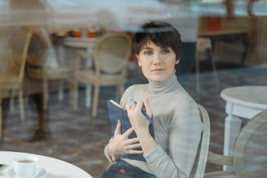 Smiling young woman in a cafe taking notes in notepad during coffee break, view through window glass