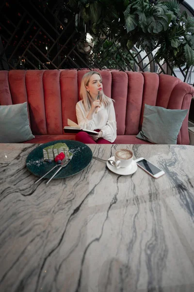 Verträumte junge Frau schreibt Zukunftspläne und Ziele in ihr eigenes Tagebuch und ruht sich in gemütlicher Cafeteria aus — Stockfoto