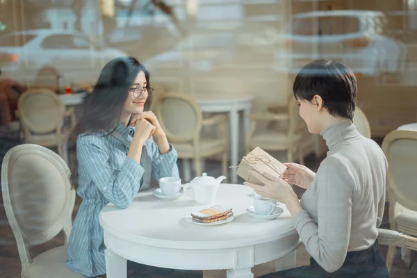 Junges schönes Mädchen sitzt mit ihrem Freund im Café und erhält ein Geschenk, Blick durch das Fenster — Stockfoto