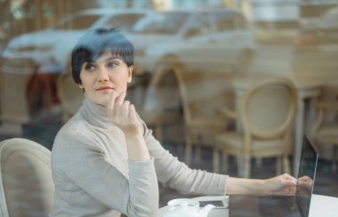 Düşünceli bir kavram. Kafede bilgisayar başında çalışan bir kadın. Pencere camından bakıyor.