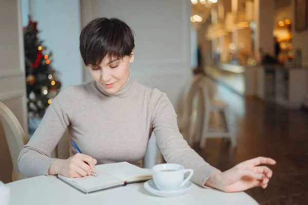 Приваблива молода жінка з ручкою в руці думає про плани і список письма, щоб зробити в блокноті, насолоджуючись відпочинком в стильному кафе — стокове фото
