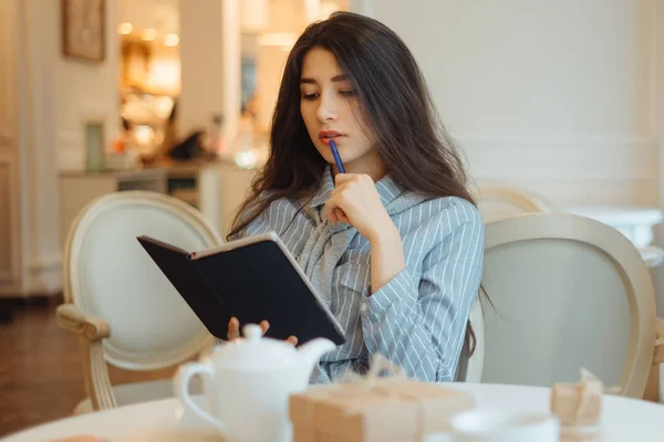 Продумана молода жінка тримає блокнот і думає про творчі ідеї для написання есе в кафе — стокове фото