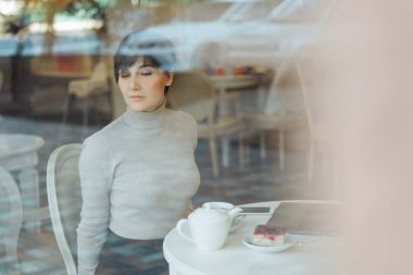 Düşünceli bir kavram. Kafede bilgisayar başında çalışan bir kadın. Pencere camından bakıyor.