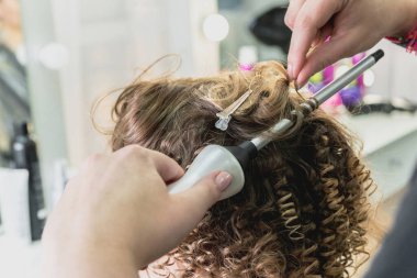 Hairdresser working with beautiful woman in hairdressing salon. Close up view of hand, curling iron and the appliance