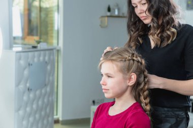 Hairdresser weaves a braid to a preteen blond girl in a beauty and hair salon