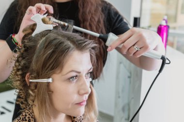 Hairdresser working with beautiful woman in hairdressing salon. Close up view of hand, curling iron and the appliance