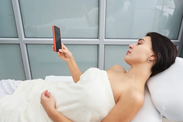 Mujer joven atractiva usando un teléfono inteligente y mensajes de texto mientras está acostado en una cama de masaje en un spa —  Fotos de Stock