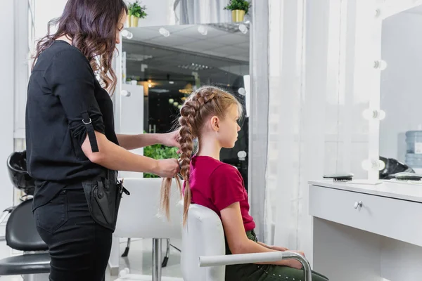Hairdresser weaves a braid to a preteen blond girl in a beauty and hair salon — Stock Photo, Image