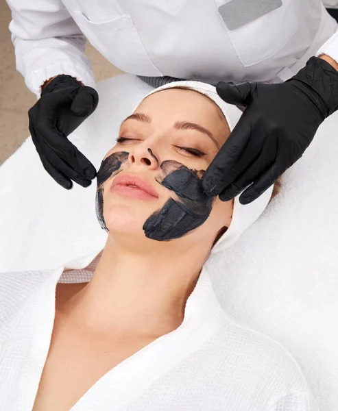Cosmetologist aplicando máscara preta no rosto de uma mulher bonita para a casca de carbono — Fotografia de Stock