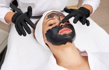 Cosmetologist applying black mask on the face of a beautiful woman for carbon peel clipart