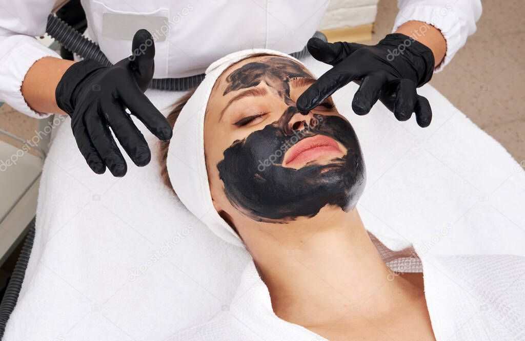 Cosmetologist applying black mask on the face of a beautiful woman for carbon peel