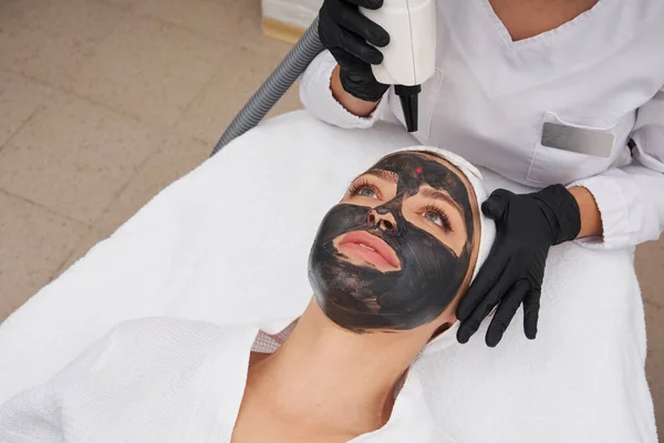 Cosmetologist aplicando máscara preta no rosto de uma mulher bonita para a casca de carbono — Fotografia de Stock