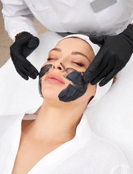 Cosmetologist aplicando máscara preta no rosto de uma mulher bonita para a casca de carbono — Fotografia de Stock