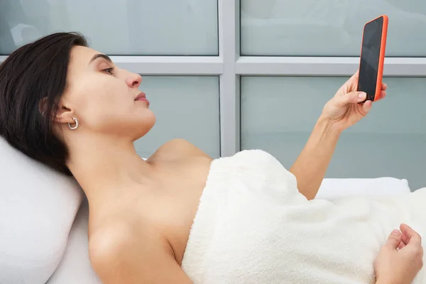 Mujer joven atractiva usando un teléfono inteligente y mensajes de texto mientras está acostado en una cama de masaje en un spa —  Fotos de Stock