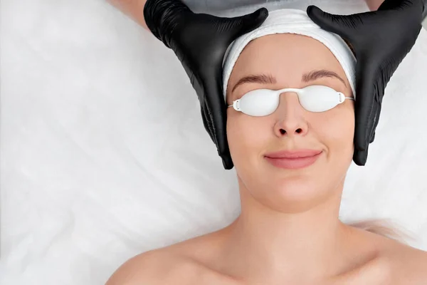 Beautiful young woman with covered eyes is getting face skin treatment. Doctor in medical gloves is going to do the procedure — ストック写真