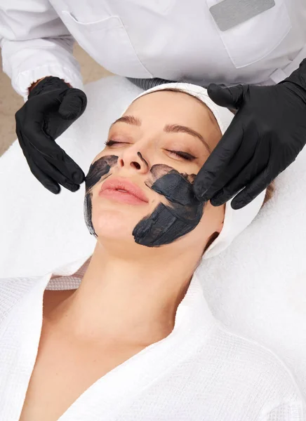 Kozmetológus fekete maszkot visel egy gyönyörű nő arcán szénhámlásra. — Stock Fotó