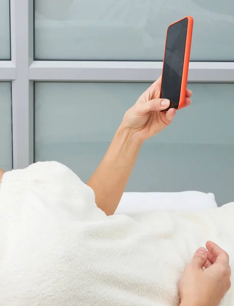 Mujer joven atractiva usando un teléfono inteligente y mensajes de texto mientras está acostado en una cama de masaje en un spa —  Fotos de Stock