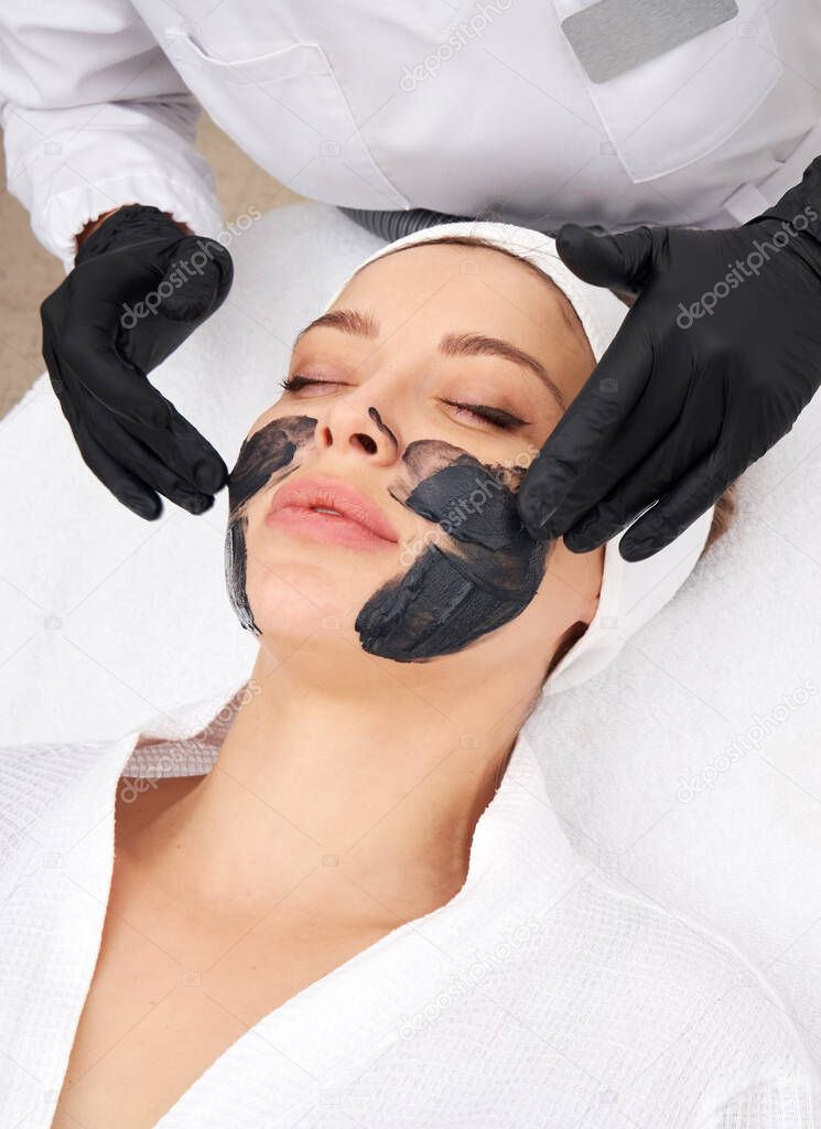 Cosmetologist applying black mask on the face of a beautiful woman for carbon peel