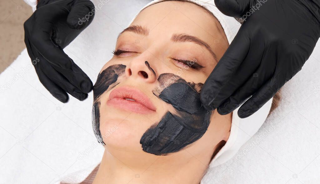 Cosmetologist applying black mask on the face of a beautiful woman for carbon peel