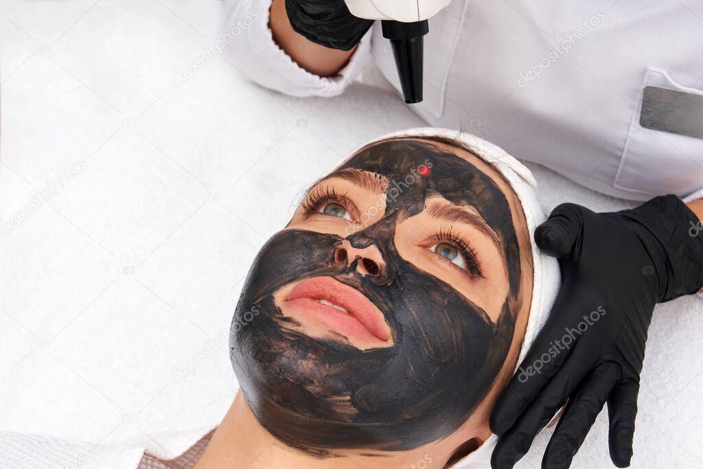 Cosmetologist applying black mask on the face of a beautiful woman for carbon peel
