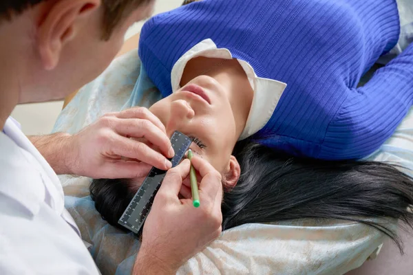 Cirurgião plástico e mulher jovem com linhas de correção antes da cirurgia estética, traçando linhas nos olhos — Fotografia de Stock