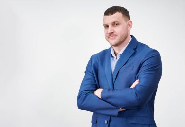 Başarılı genç iş adamı, çapraz kollu, mavi takım elbiseli, güzel, samimi bir gülümseme takınarak gri arka planda fotokopi alanı ile poz veriyor.