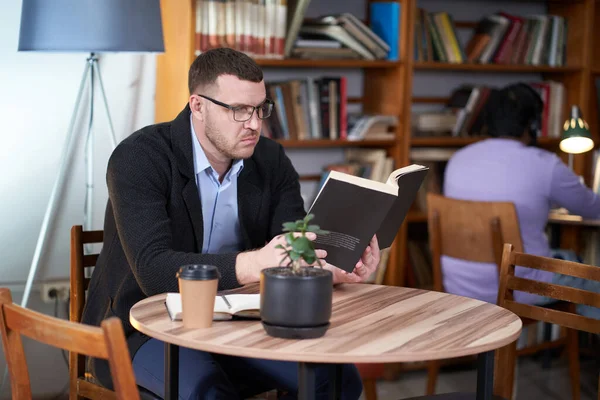 Hombre leyendo libro y bebiendo café en un café con muchos libros en un fondo — Foto de Stock