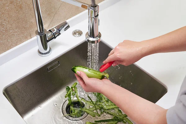 Vista recortada de manos femeninas pelando pepino sobre la máquina de eliminación de residuos de alimentos en el fregadero en la cocina moderna — Foto de Stock