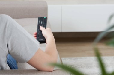 Televizyon izleyen ve elinde uzaktan kumandayla kanepede oturan genç bir kadın.