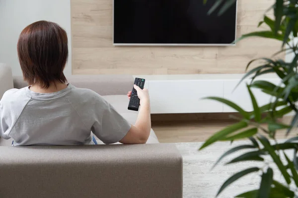 Вид спереду молода жінка дивиться телевізор і тримає пульт дистанційного керування, сидячи на дивані — стокове фото