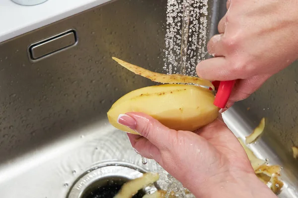 Обрезанный вид женских рук, очищающих картофель над машиной по утилизации пищевых отходов в раковине на современной кухне — стоковое фото